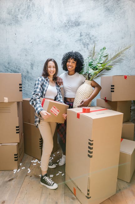 A woman with curly black hair and a bright smile is sitting on a wooden floor inside a residential space, surrounded by numerous medium-sized cardboard moving boxes, some sealed with red packing tape.